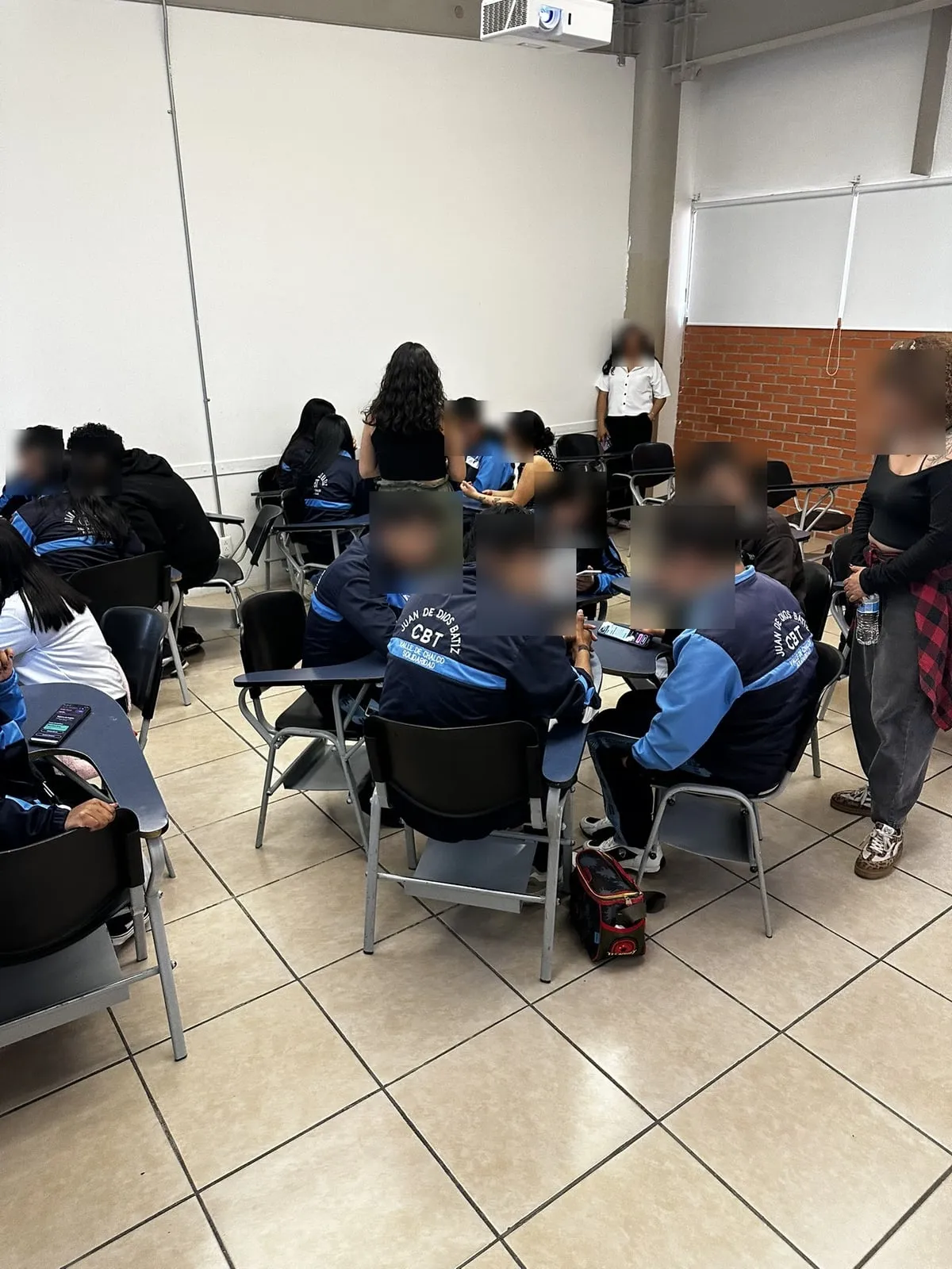 Un equipo de estudiantes con uniforme reunido alrededor de un teléfono; una facilitadora observa de pie. Los rostros están difuminados.