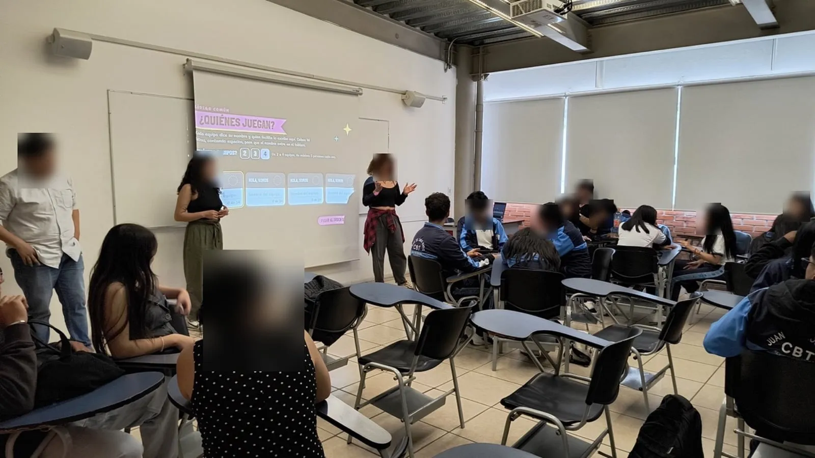Dos facilitadoras explican el juego junto a la proyección mientras los equipos escuchan desde sus lugares; los rostros están difuminados.