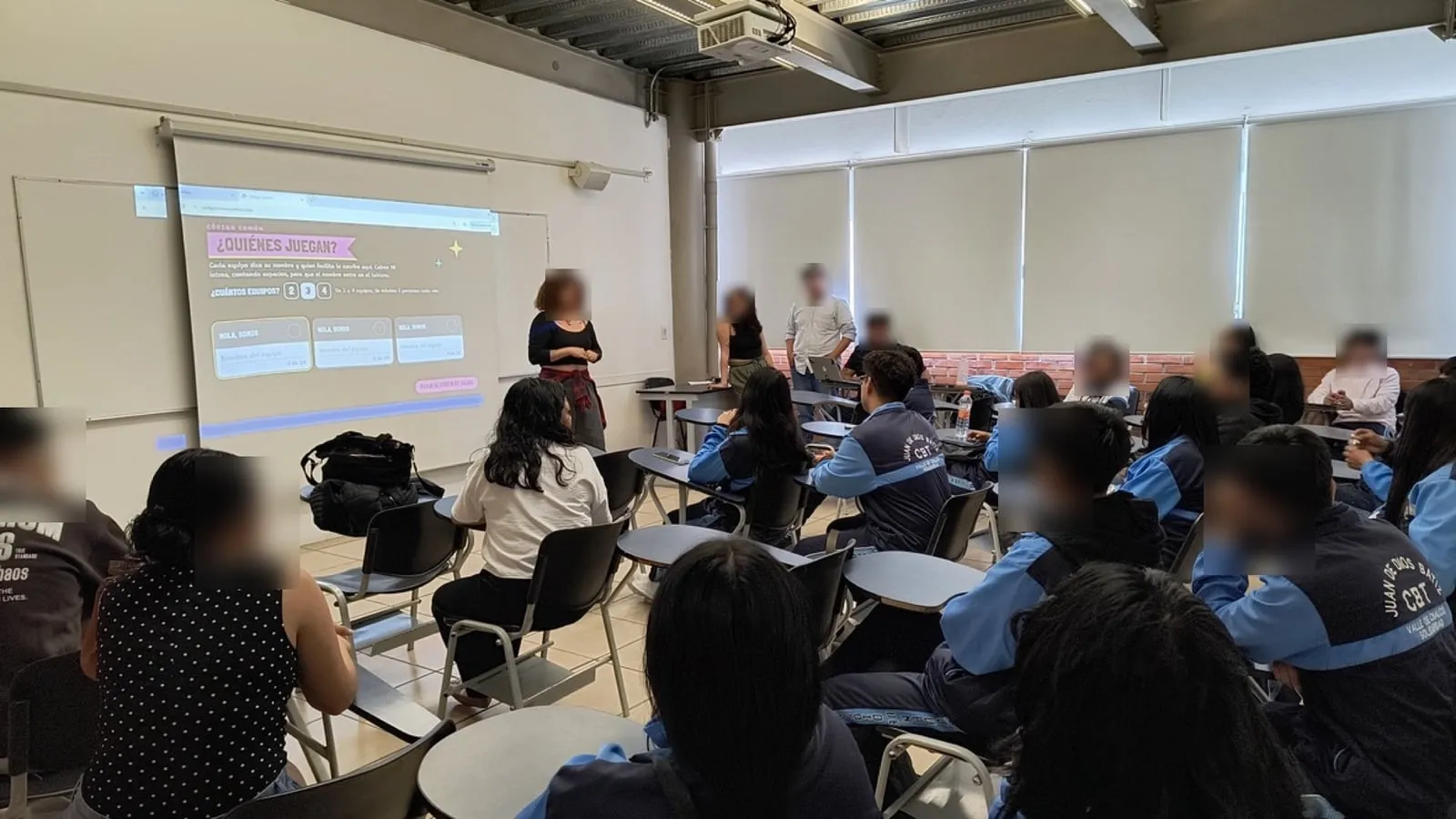 Salón con estudiantes sentados frente a la proyección de Código Común, que pregunta «¿Quiénes juegan?»; los rostros están difuminados.