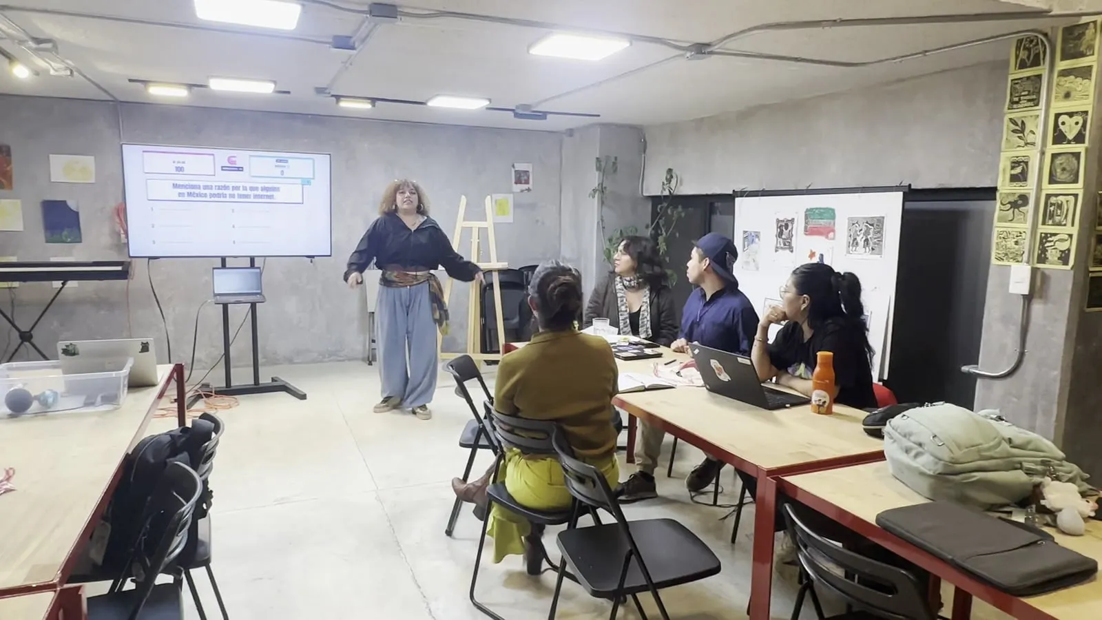 Sala con mesas de trabajo; al fondo, una TV muestra el tablero de Esto respondieron y una persona conduce de pie.